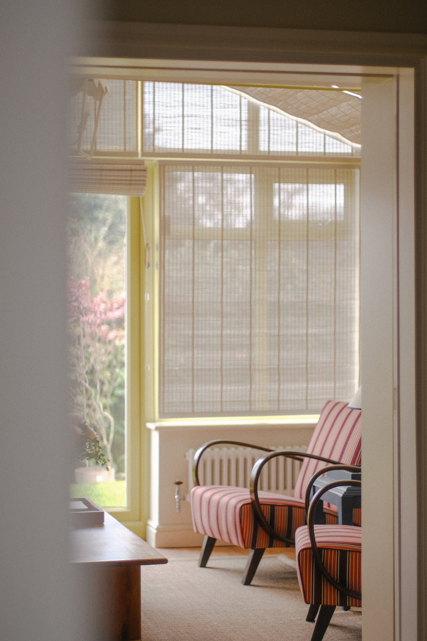 Striped upholstered armchairs positioned beside glazed doors and woven blinds in a light-filled garden room.