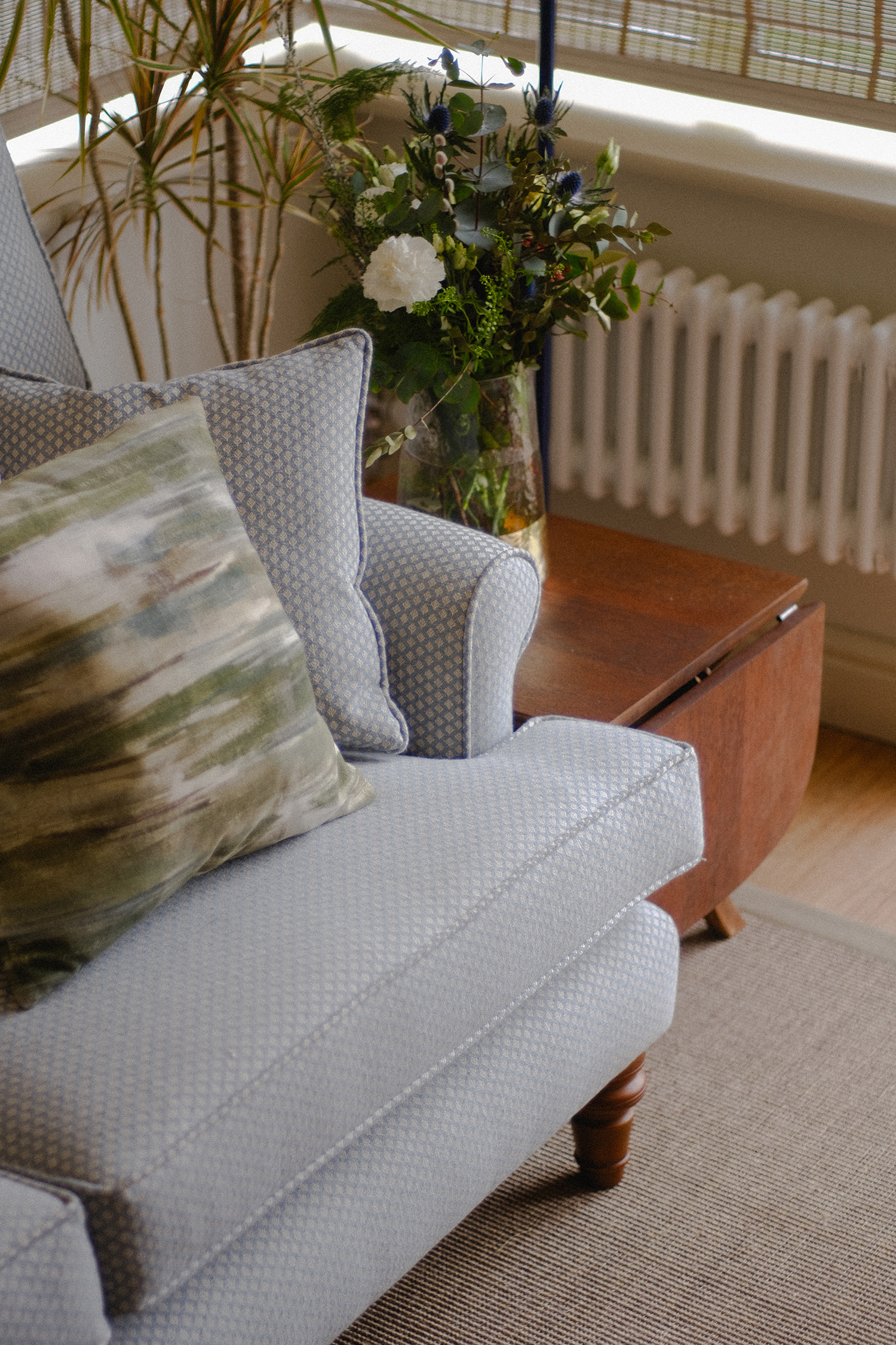 Close-up of sofa arm, timber side table and fresh floral arrangement beside a traditional radiator.