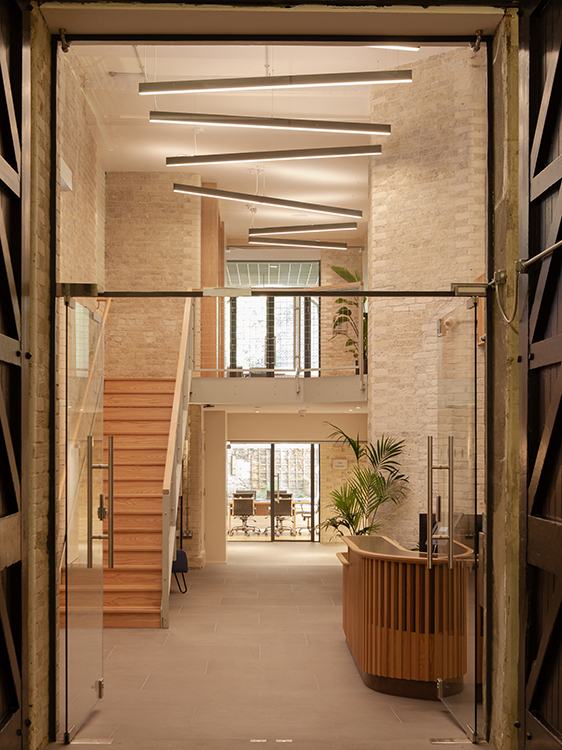 Reception space within a Clerkenwell workplace retrofit featuring exposed brickwork, bespoke timber reception desk, new staircase and retained industrial structure.