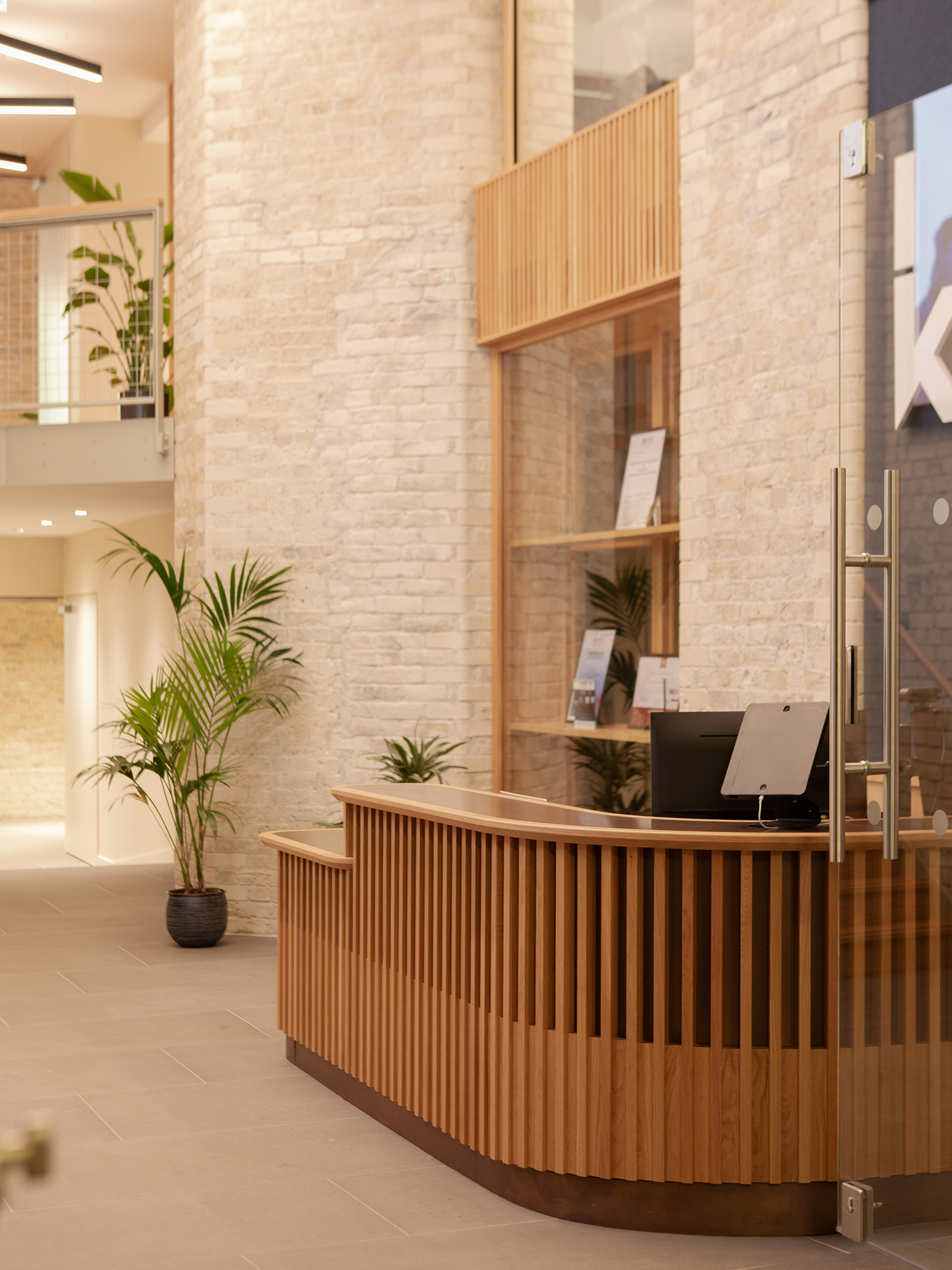 Reception desk with a curved timber slatted front in a bright, double-height interior with pale brick walls and indoor plants