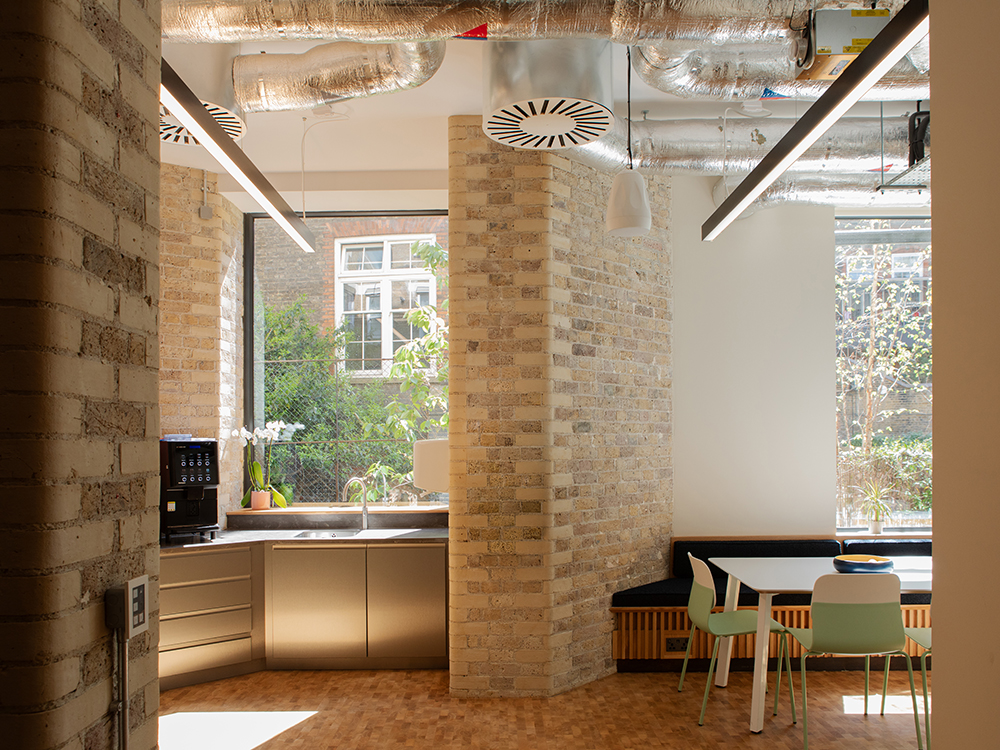 Breakout and kitchen area with exposed brickwork, timber flooring, integrated seating and views to a landscaped courtyard.