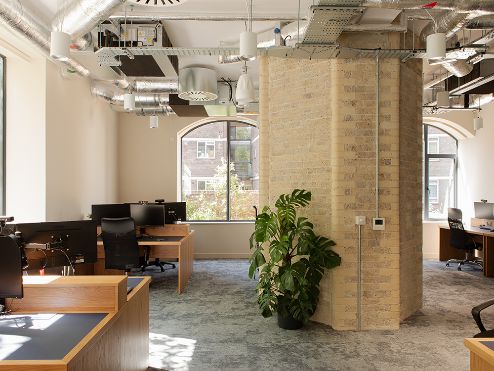 Open-plan office with retained brick columns, bespoke timber workstations, exposed services and natural daylight.