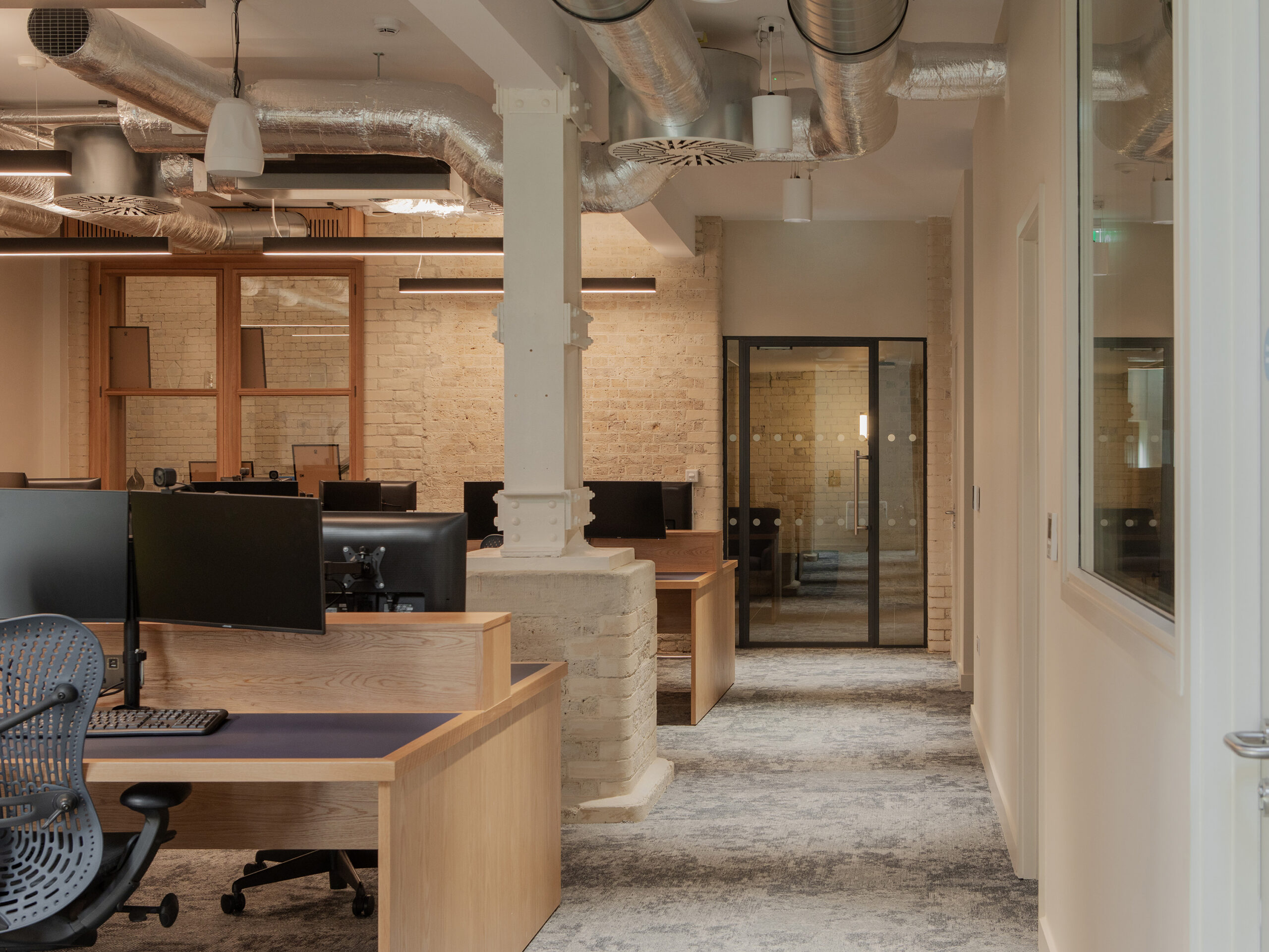 Open-plan office within a retained industrial building featuring exposed brickwork, bespoke timber workstations, original structural columns and glazed meeting rooms.