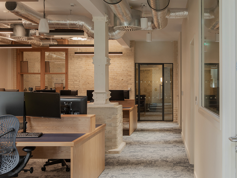 Open-plan office within a retained industrial building featuring exposed brickwork, bespoke timber workstations, original structural columns and glazed meeting rooms.
