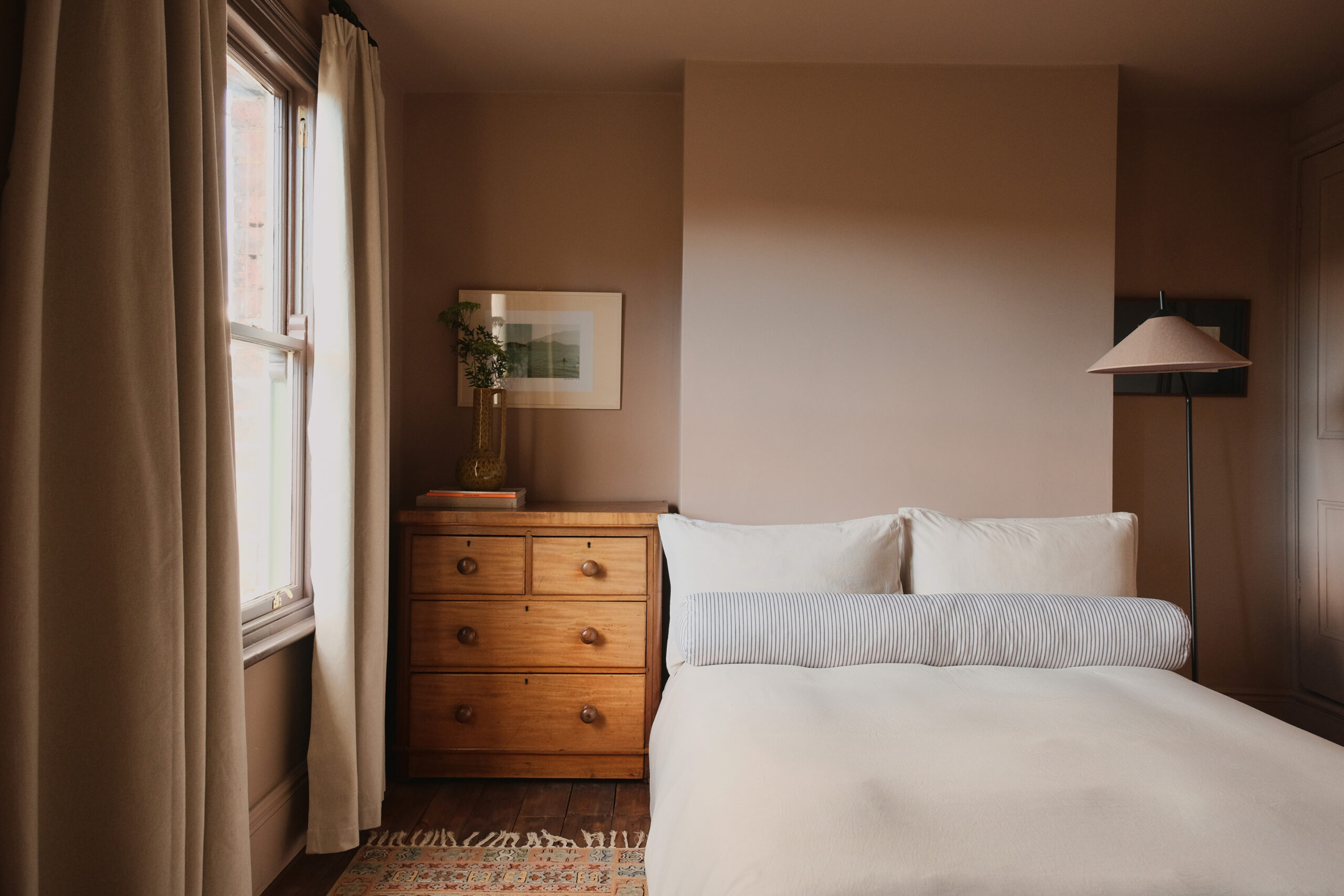 Bedroom with warm plaster-toned walls, timber furniture, linen curtains and original floorboards.
