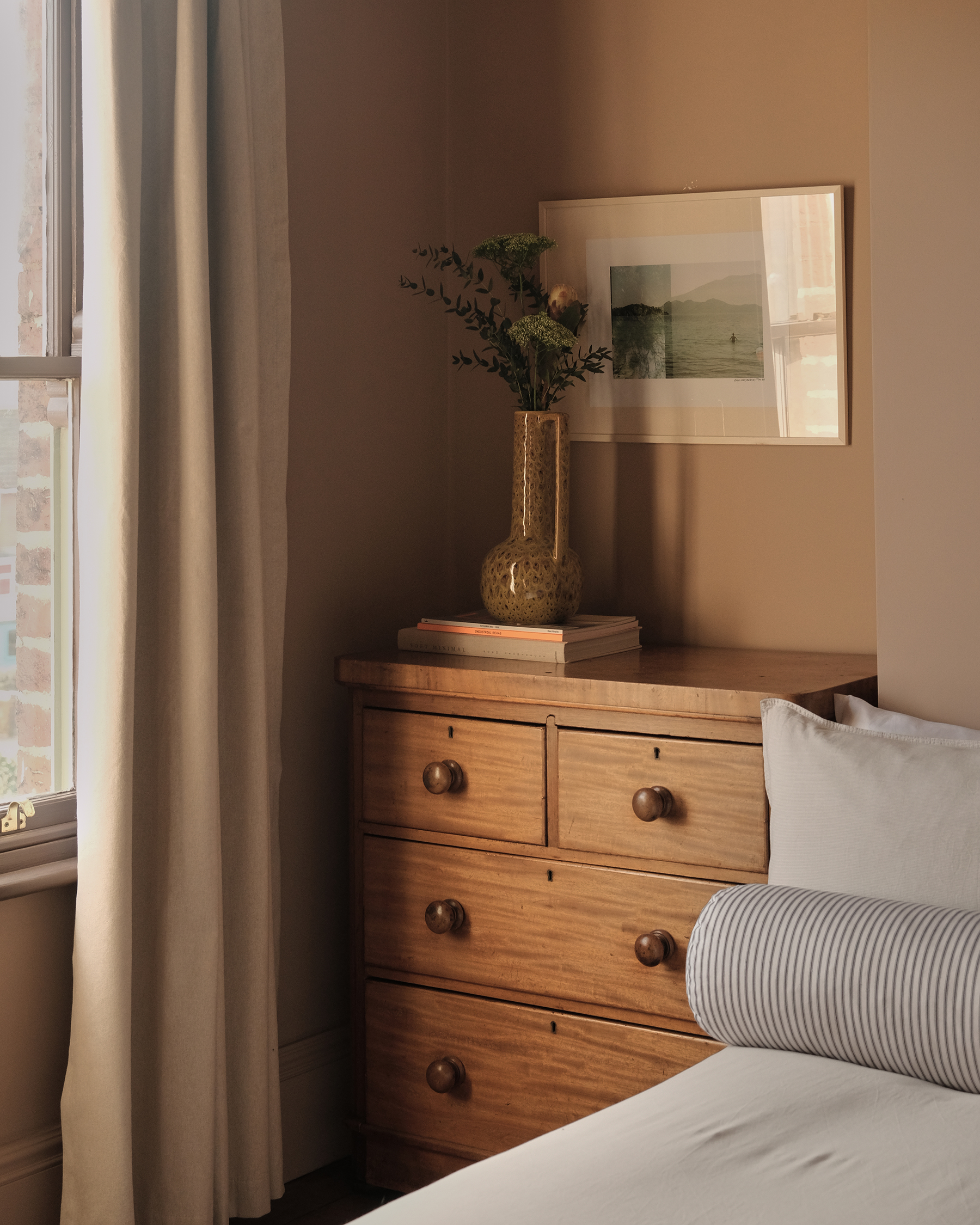 Wooden chest of drawers beside a bedroom window, with a vase, framed artwork and the edge of a bed
